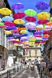Colourful Umbrellas Hanging In The Air As Decorations Above A Shopping Street In Arles With Shady Shadows Cast Onto Road