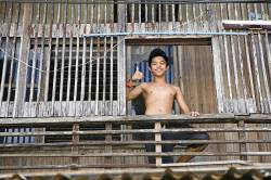 A Filipino Teen Boy Stands On The Deck Of His Traditional Philippine Style Wooden Home , Bahay , And With A Smile Gives A Friendly Thumbs Up .