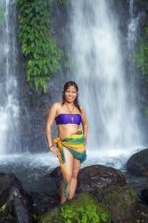 A Beautiful, Sexy Asian Woman Wearing A Colourful Wrap And Blue Halter Stands In Front Of A Tropical Waterfall In Southeast Asia.