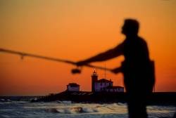 Silhouette Of A Man Surfcasting At Misquamicut State Beach, Rhode Island, USA In Late Afternoon Against Deep Sunset Colors. Watch Hill Lighthouse, Westerly, Rhode Island, USA In The Background.