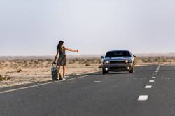 Woman Hitch Hiking On Rural Road In The Desert With Her Luggage And A Car Coming Behind Her