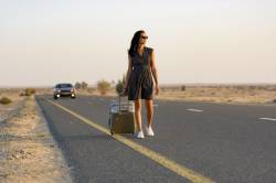 Woman Walking With Luggage On Rural Road In The Desert With A Car Coming Behind Her